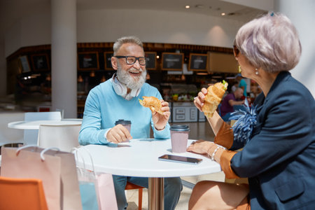 Overjoyed retired couple eating croissant at cafeteria in shopping mallの写真素材