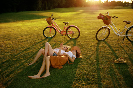 Female friends sitting on grass laughing and talkingの写真素材
