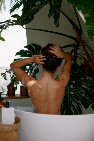 View from back on beautiful woman massaging neck while taking bath at homeの写真素材