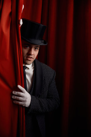 Handsome man magician peeking out from behind stage drapery curtain