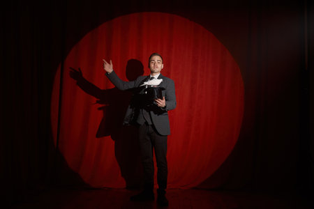 Magician with pigeon standing over red drapery in spotlight