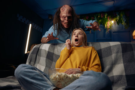 Shocked woman watching movie with popcorn. Man wearing zombie mask behind wants to scary her