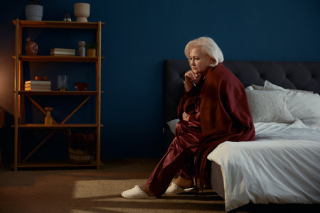 Elderly woman wearing pajamas sitting on bed feeling lonelinessの写真素材