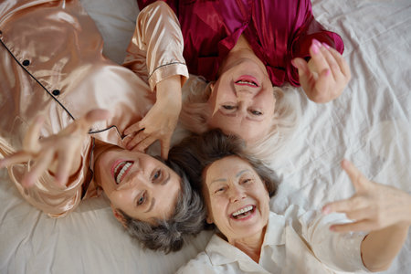 Top view on smiling elderly woman friends wearing pajamas lying in bedの写真素材