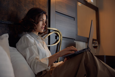 Businesswoman working online while resting in bed at hotel roomの写真素材