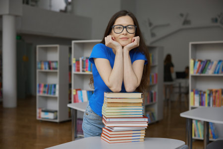 Young woman overloaded with huge stack of paper book standing over bookstoreの写真素材