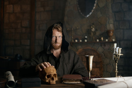 Mystical spooky medieval monk in black cassock with human skull on table
