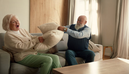 Playful elderly couple fighting with pillows having fun time togetherの写真素材
