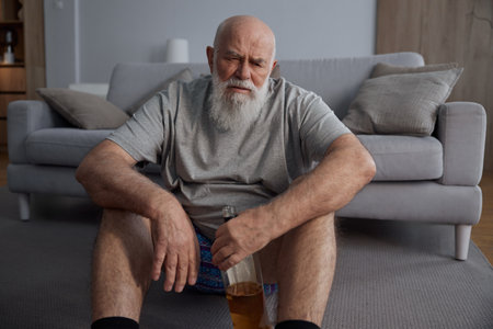Senior man drinking alcohol alone sitting on floor at homeの写真素材