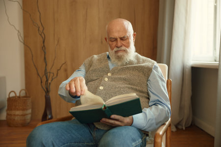 Serious retired senior man reading paper book sitting in cozy armchair at homeの写真素材
