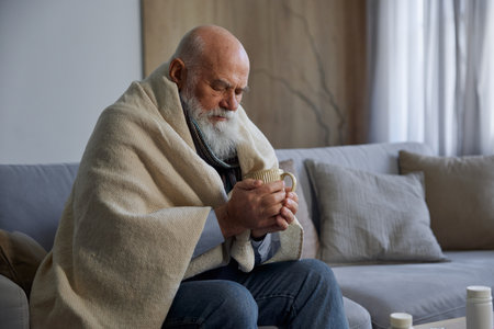 Sick weak senior man feeling unwell drinking tea sitting on sofaの写真素材