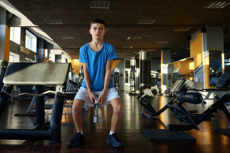 Athletic teenage boy squatting with dumbbells training over gym interiorの写真素材