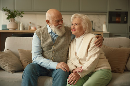 Portrait of happy elderly married couple sitting on sofa in living roomの写真素材