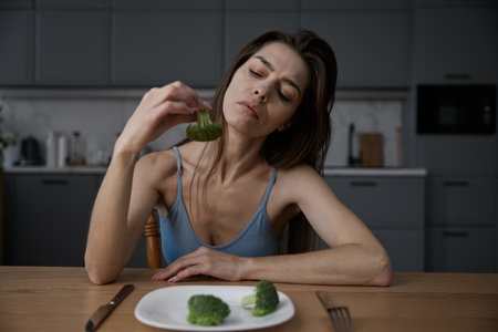 Displeased woman suffering from eating disorder eating broccoliの写真素材