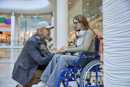 Lovely grandfather helping his granddaughter who sits in a wheelchair in a shopping mallの写真素材