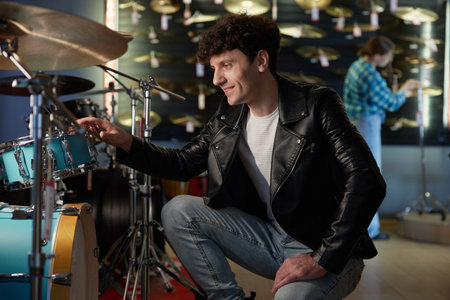A man wearing a stylish leather jacket is sitting upon a drum set