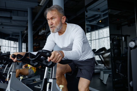 Elderly man riding an exercise bike in a bustling gymの写真素材