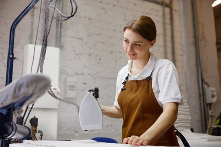 A woman worker wearing an apron is skillfully ironing a shirt on a tableの写真素材