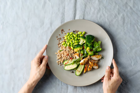Healthy eating concept. Woman hands holding plate with brown rice, broccoli, chicken fillet and cucumber on gray background, top viewの素材