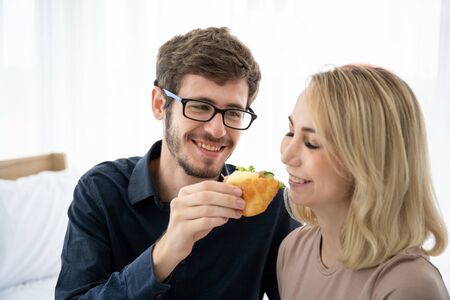Caucasian young couple portrait in bedroom concept. Boyfriend embracing and feeding his girlfriend a piece of sandwich in bed room in morning. Passionate love of young couple having romantic moment.の写真素材