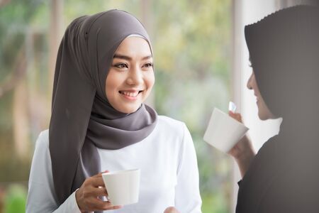 Two Asian Muslim women standing and talking in the office with a cup of coffee. Modern Muslim...