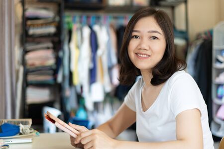 Beautiful young adult Asian designer in a boutique is using a touchscreen tablet to design a new clothing. Small Asian's clothing merchandise business concept. Girl portrait in clothing room.の写真素材