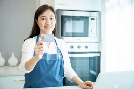 Happy Asian woman presenter showing a credit card while using notebook in a kitchen, girl make an online shopping on laptop computer and pay via credit card. Online transaction and payment concept.の写真素材