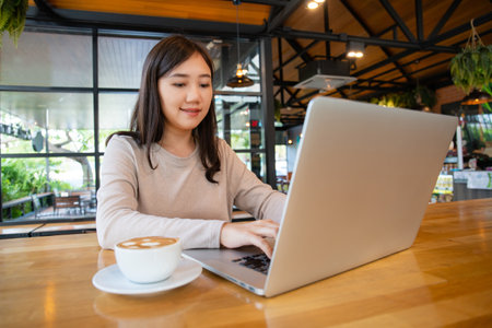 Asian beautiful working woman doing her work and drink in the coffee shop or cafe. Cheerful online blogger writing on her laptop or notebook computer and drinking a coffee close up. Modern lifestyle.の写真素材