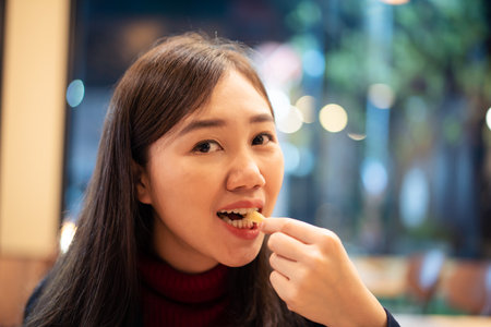 Asian woman enjoy eating French fry or potato chip in the fast food restaurant at night. Unhealthy eating behavior in young adult. Often eating the junk food is not good for health.の写真素材