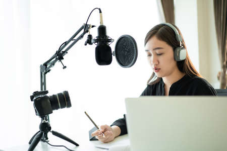 Happy Asian woman setting up a living room in her house for podcast studio, woman arranging a podcast and online radio station at home. Professional young podcaster speaking through a microphone.の写真素材