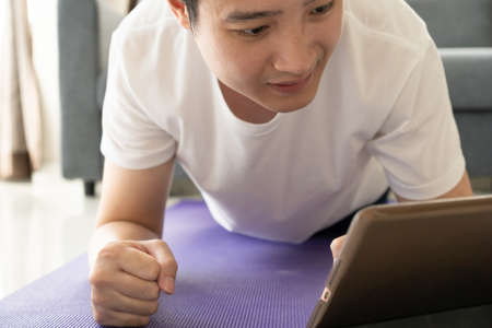 Young Asian young man doing a plank and push up exercise and watching on a tablet, tablet isolated screen in white with clipping path. Man learning and training an exercise online on tablet.の写真素材