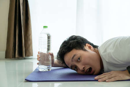 Funny tired face of Asian young man laying down on the floor and trying to grab a water in bottle. Man tired from hard workout.の写真素材