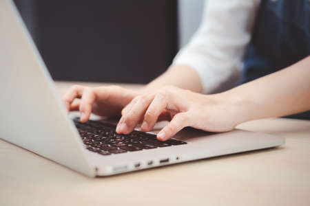 Unrecognizable Asian young woman using laptop computer working on the wooden table in restaurant -...
