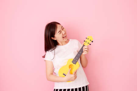 Happy Asian woman holds acoustic Ukulele guitar on pastel color background close up with copyspace. Cheerful young girl playing and practicing a Hawaiian guitar. Female hand holding the Ukulele.の写真素材