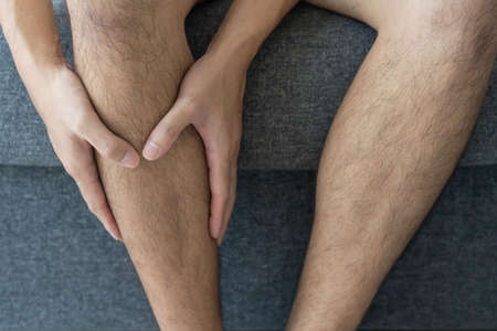 Unrecognizable Asian young man sitting on the sofa in living room and touching at his leg, man got an injured at leg.の写真素材