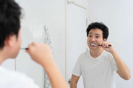 Happy Asian young man in white t-shirt brushing a teeth in bathroom in the morning. A man preparing...