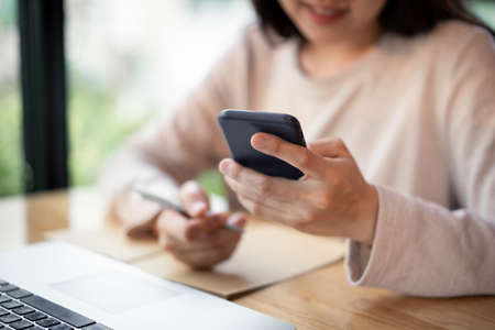 Unrecognizable cheerful young businesswoman reading a message on a smartphone close up. Asian girl working on a laptop computer and smartphone during staying at coffee shop. Online learning concept.の写真素材