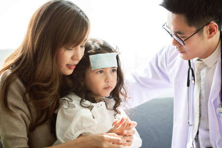 Asian Mother takes her sick young daughter to see a doctor in hospital and doctor pasting a cool fever gel on girl's forehead, girl got a sickness from flu virus. Young girl patient got a sick.の写真素材