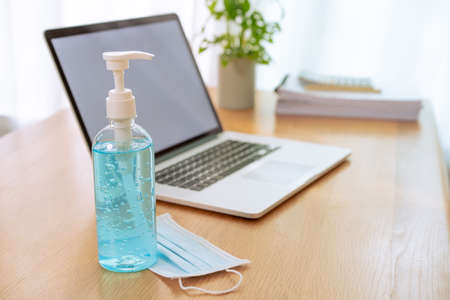 Liquid hand sanitizer, medical hygiene protective face mask and laptop computer left on the wooden table in the hospital office, COVID-19 or Coronavirus pandemic awareness with new normal lifestyles.の写真素材