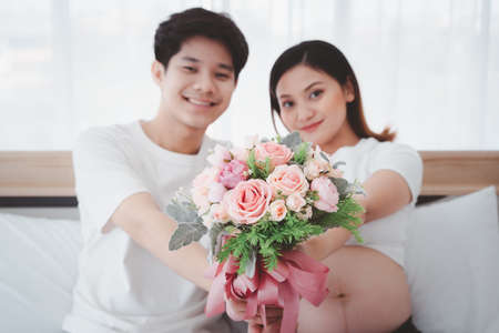 Cheerful Asian family, husband surprise his pregnant wife with a beautiful bouquet of flower close up. Pregnancy wife stay with her lovely husband in bedroom.の写真素材