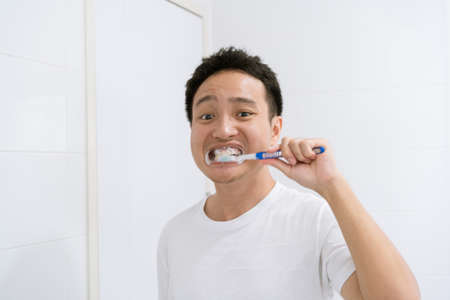Asian young man in white shirt brushing his tooth in front of the mirror in the morning after got up from the bed, oral health care and wellbeing. Dental care concept.の写真素材