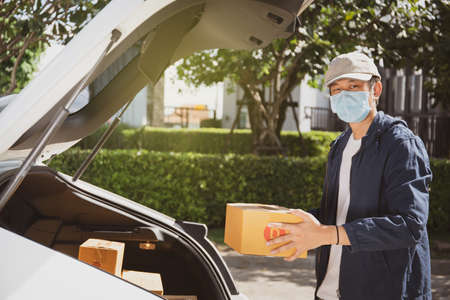 Asian postman with a hygiene protective face mask picking up a parcel package or parcel box from the truck for deliver to customer's door. New Normal concept during covid-19 or coronavirus pandemic.の写真素材