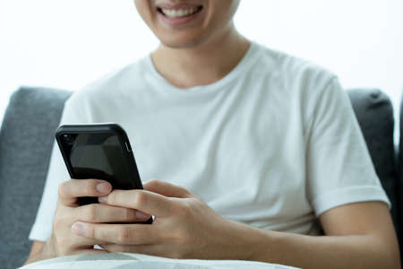 Asian young man in white t-shirt using a touchscreen smartphone while staying in the living room...