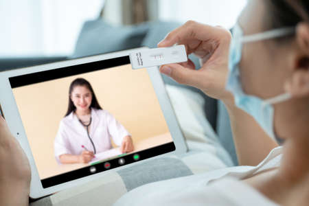 Asian young woman with hygiene protective face mask using SARS 2019-nCoV COVID-19 coronavirus antigen rapid test kit - ag test kit at home and consulting a testing result with doctor via video call.の写真素材
