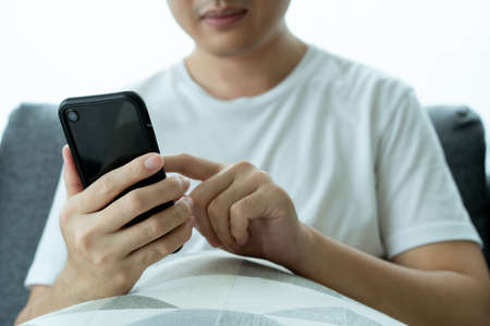 Asian young man in white t-shirt using a touchscreen smartphone while staying in the living room close up. Man browsing the internet and social media on his smartphone.の写真素材