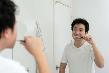 Happy Asian young man in white t-shirt brushing a teeth in bathroom in the morning. A man preparing and checking his face and body before go to work. Routine activity in daily life.の写真素材
