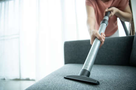 Asian housewife using a wireless vacuum machine to clean a sofa in living room close up. Asian housekeeper vacuuming in living room. Chores and daily housework activities concept.の写真素材