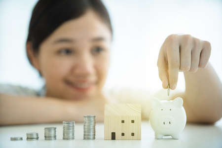 Happy beautiful Asian young woman putting a coin into saving piggy in concept of money saving for housing - asset. Girl saving her money for buying a house in future.の写真素材