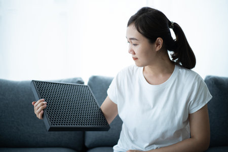 Beautiful Asian young woman holding a HEPA carbon air purifier filter in living room, woman prepare a air purifier filter to be replace the old one. Healthcare and good living lifestyle concept.の写真素材