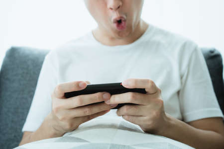 Asian young man in white t-shirt using a touchscreen smartphone while staying in the living room close up. Man browsing the internet and social media on his smartphone.の写真素材